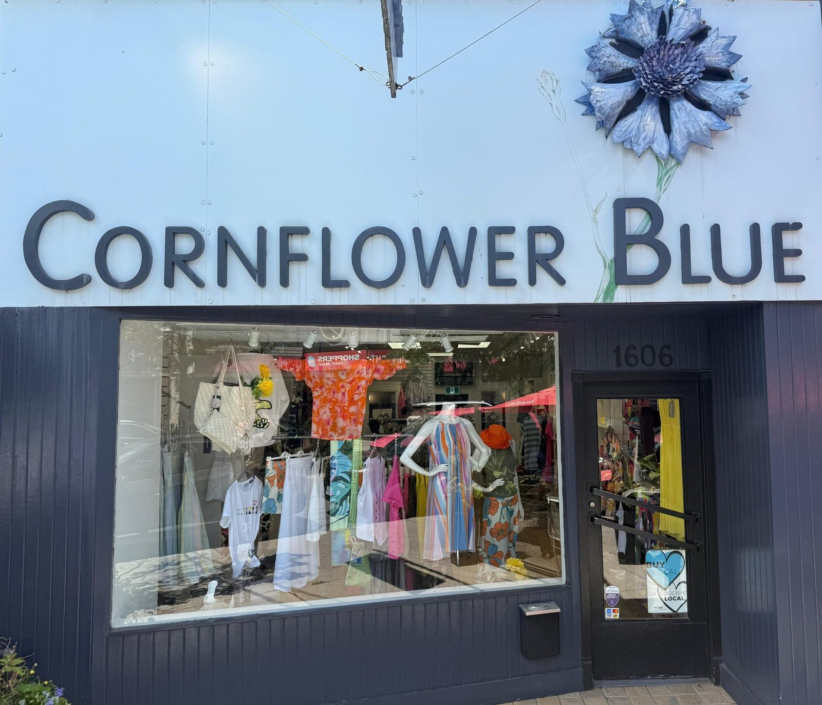 A storefront of Cornflower Blue with large lettering, showcasing colorful clothing items in the window. The display includes dresses and accessories, situated on a busy street in Toronto, highlighting local women’s fashion. women’s boutique Toronto on Bayview