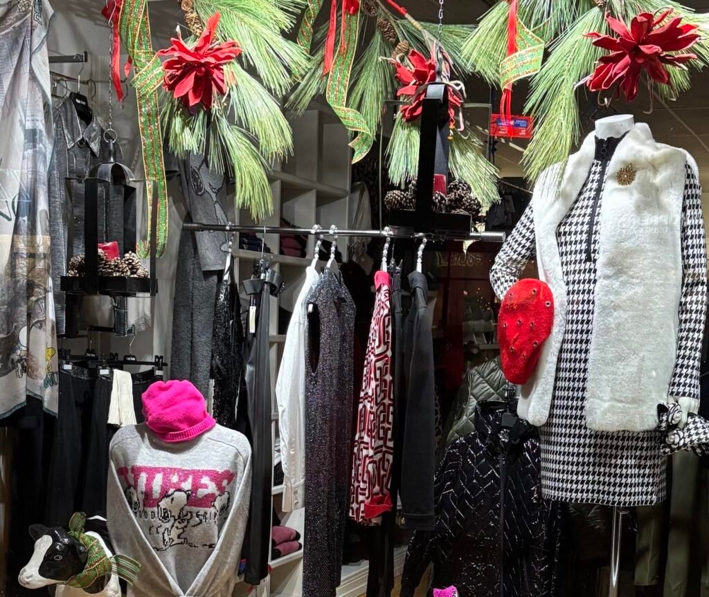 A clothing store display featuring a variety of women's apparel, including sweaters, pants, and a black and white houndstooth dress. Seasonal decorations with pine branches and red flowers are visible in the background. women’s boutique Toronto on Bayview